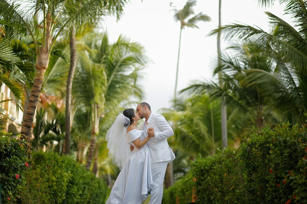 Bride and groom in white attire embracing in a tropical garden lined with palm trees at a destination wedding venue.