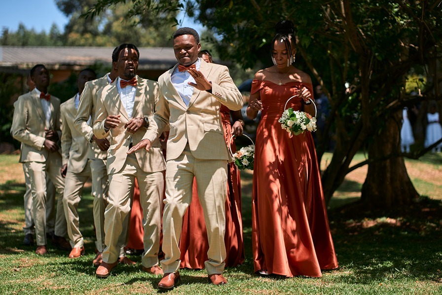 Groomsmen and bridesmaid walking outdoors in coordinated beige suits and rust dresses during a wedding celebration.
