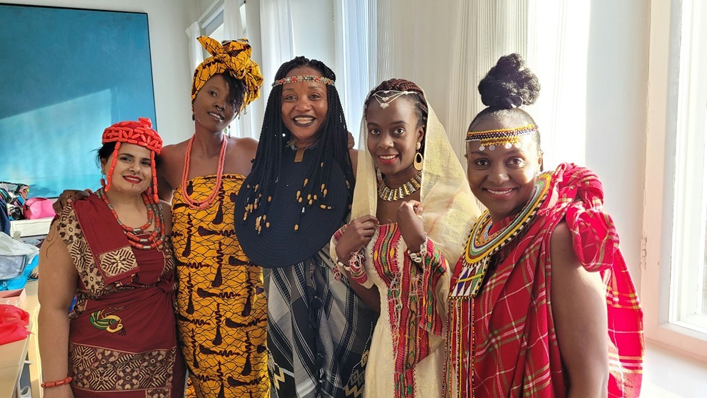 Group of women wearing colourful traditional African attire and accessories, smiling indoors during a cultural celebration.