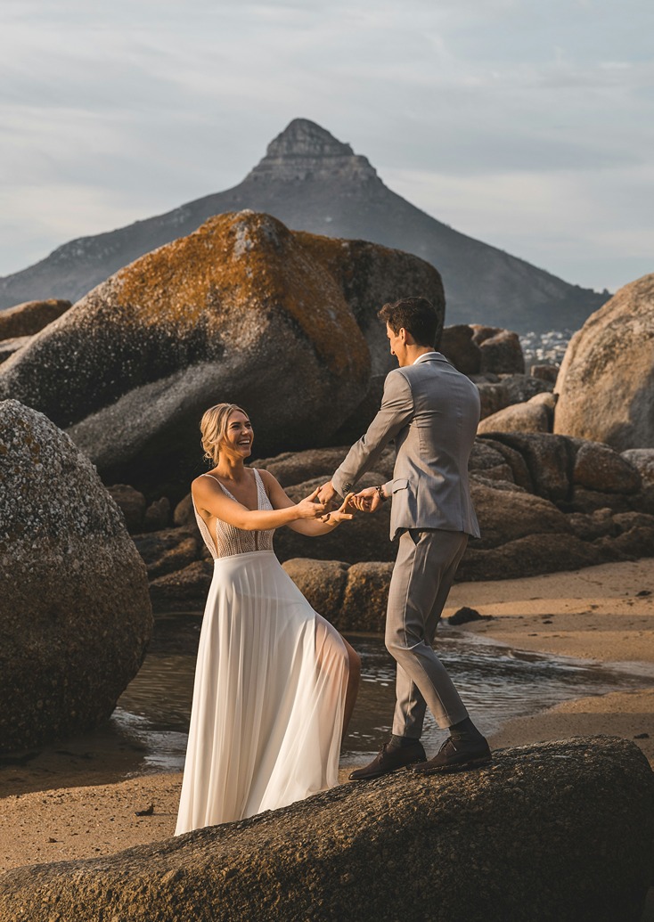 Alt text: A laughing bride and groom holding hands on the granite boulders at Camps Bay beach, with Lion's Head mountain rising in the background during golden hour.