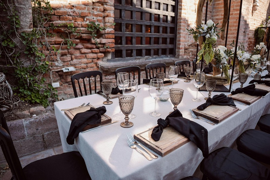 A wedding reception table set with wooden charger plates, black linen napkins, smoked glass goblets, and white floral arrangements against an exposed brick wall with climbing ivy.