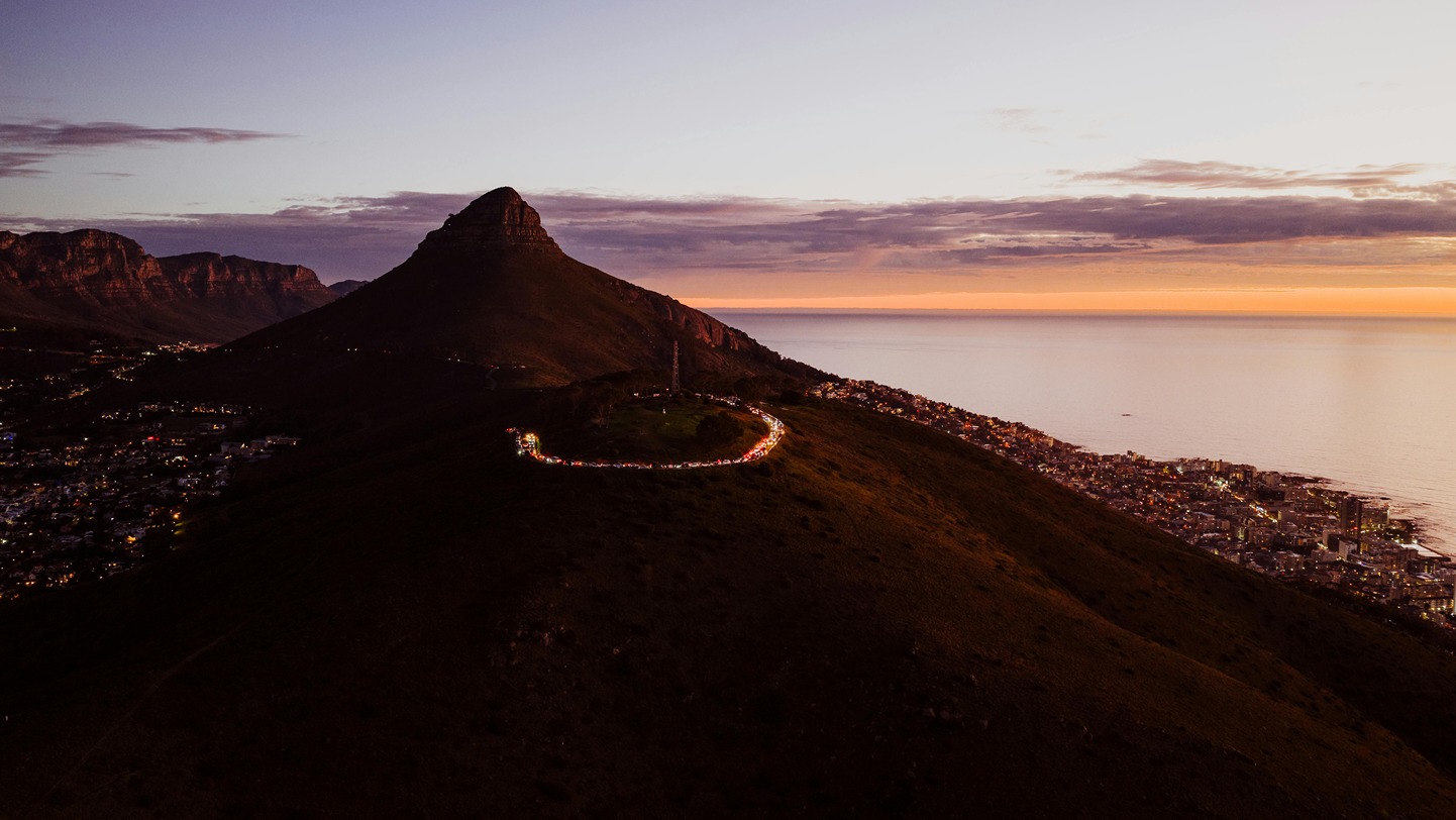 Aerial view of Lion's Head at dusk with Signal Hill in the foreground, city lights beginning to glow across Sea Point and Camps Bay, and the Atlantic Ocean reflecting the orange and purple sunset.