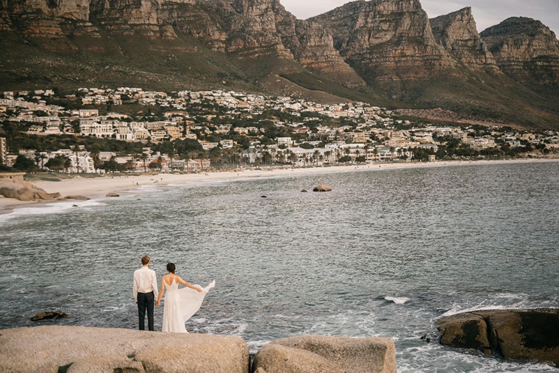 A bride and groom standing on coastal rocks holding hands, looking out over the ocean toward Camps Bay beach with the Twelve Apostles mountain range in the background.