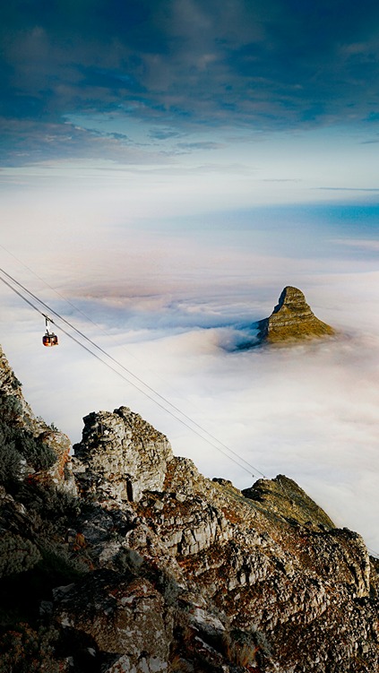 The Table Mountain Aerial Cableway descending through cables from the rocky summit, with Lion's Head peak protruding through a thick blanket of cloud below.