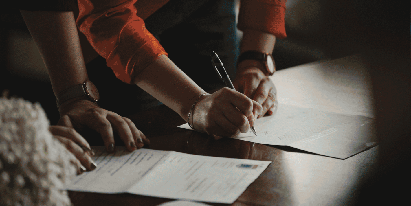 Close-up of a couple's hands signing marriage paperwork at a wooden table, with a colourful abstract border framing the photo.
